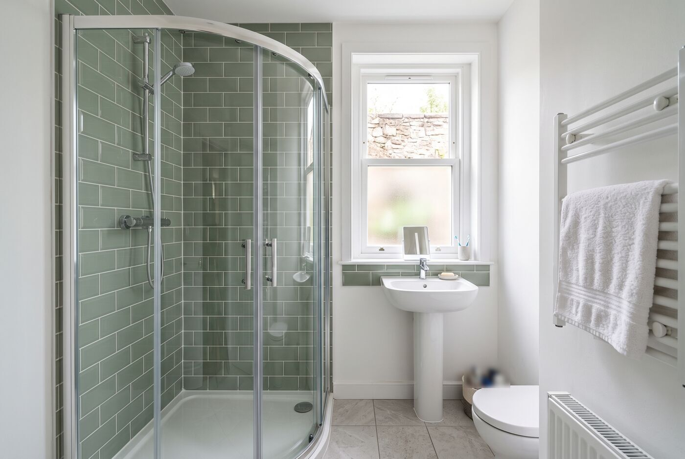 Corner quadrant shower enclosure with sage green tiles beside a pedestal basin